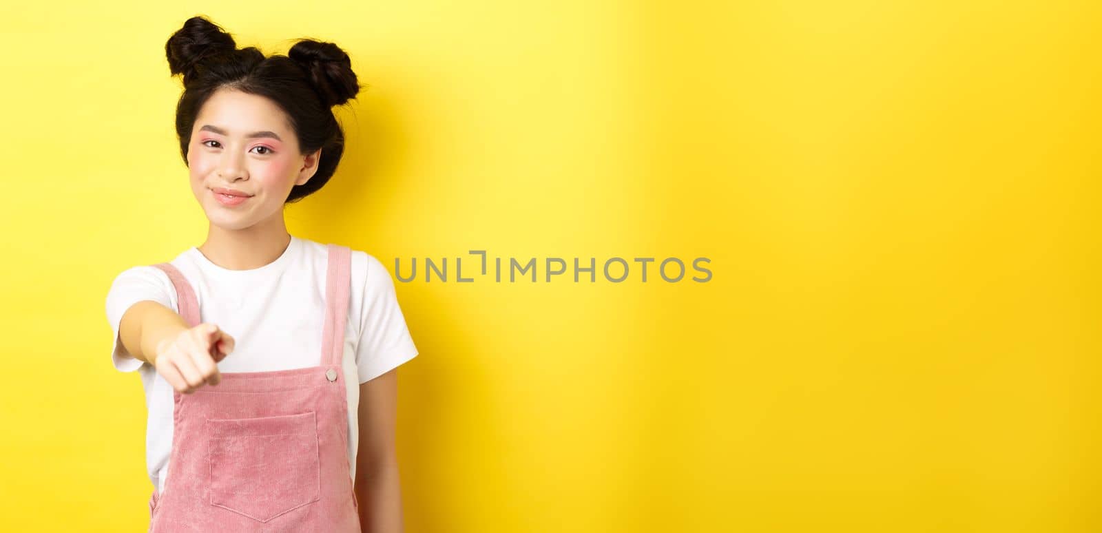 Stylish japanese girl with beauty makeup pointing at camera, smiling at you, beckon and invite people, standing on yellow background.