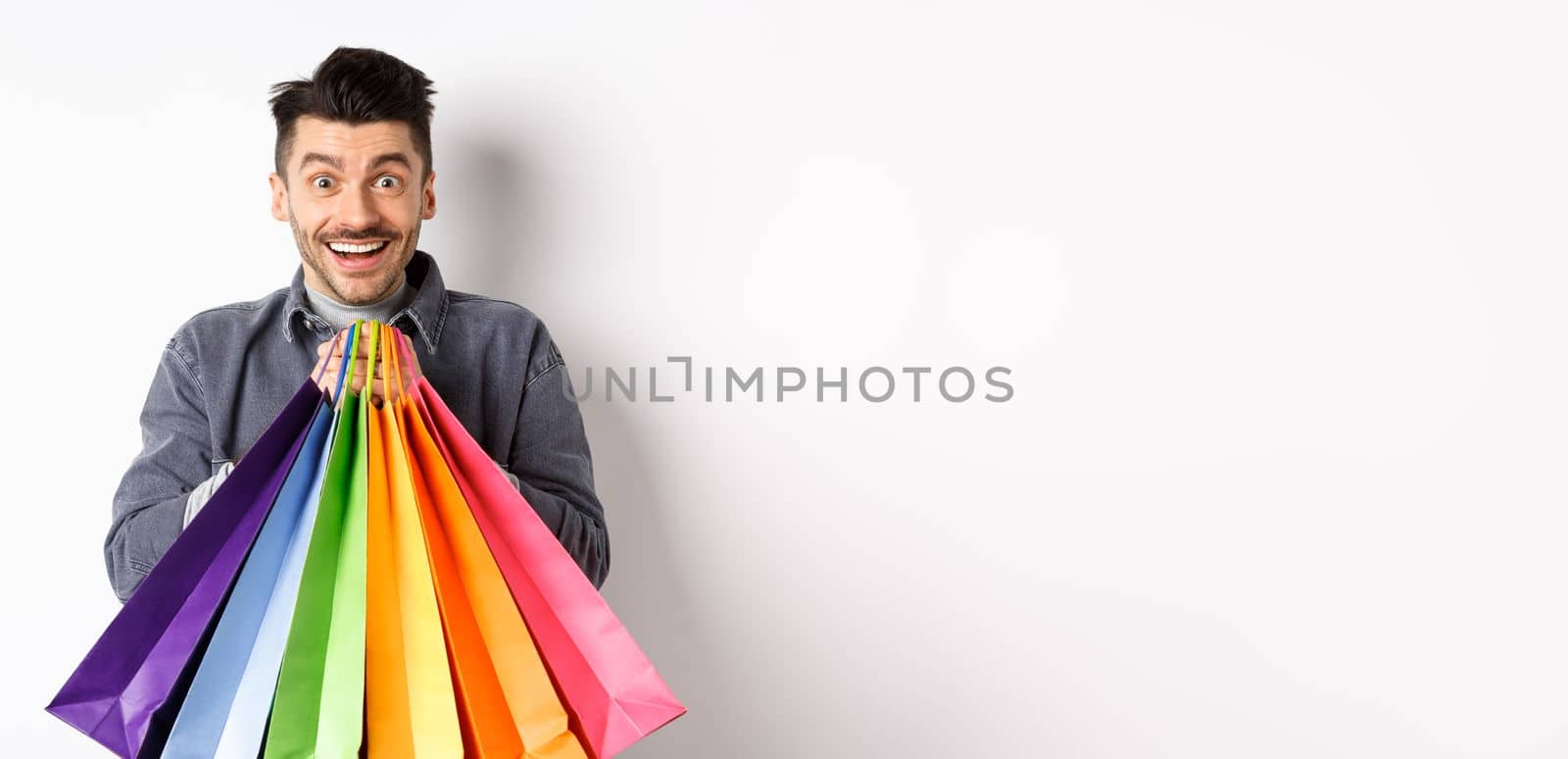 Excited smiling guy holding colorful shopping bags and rejoicing with discounts in store, standing against white background.