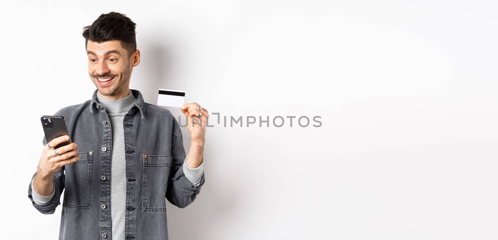 Online shopping concept. Smiling guy paying on mobile phone, showing plastic credit card, buying in internet, white background.