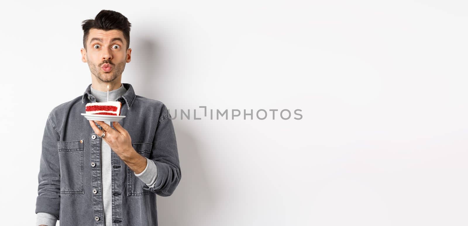 Man celebrating birthday, blowing candle on cake and making wish, standing on white background.