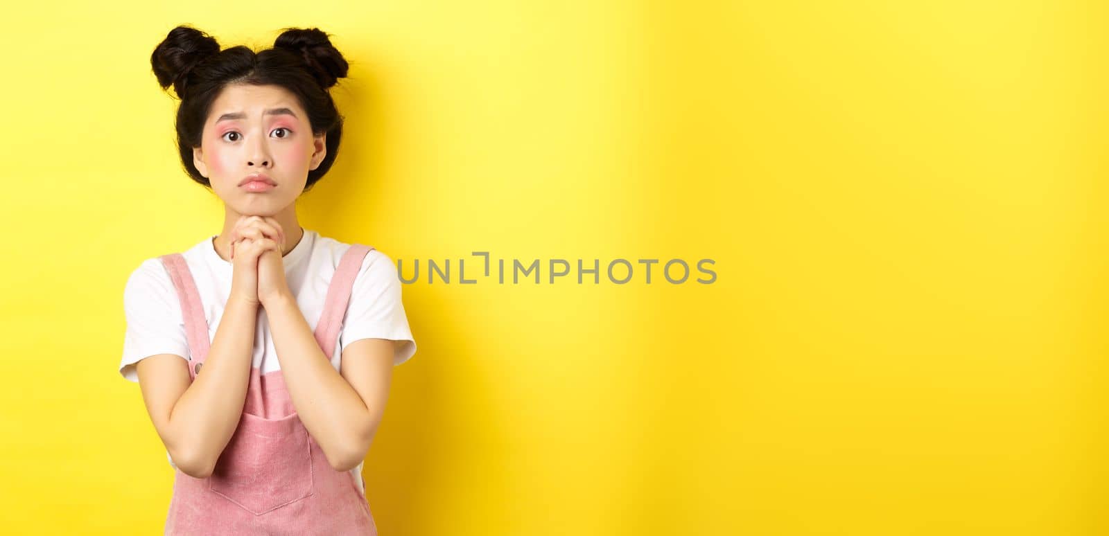 Hopeful asian stylish girl holding hands in begging sign, waiting with hope, asking for favour or help, standing on yellow background.