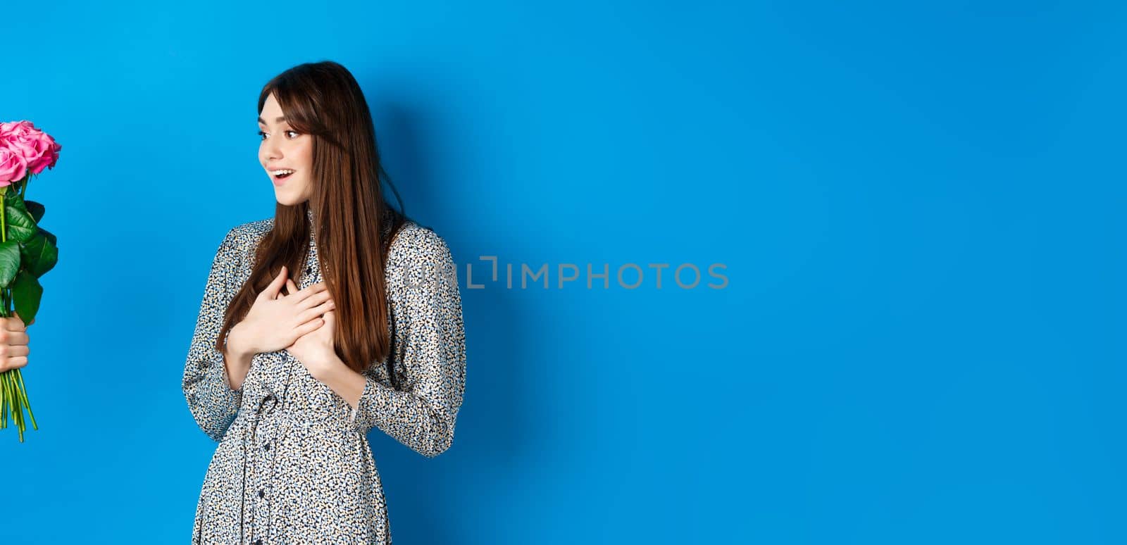 Valentines day concept. Image of surprised beautiful girl looking at hand with bouquet of flowers, receive surprise roses from lover, standing on blue background.