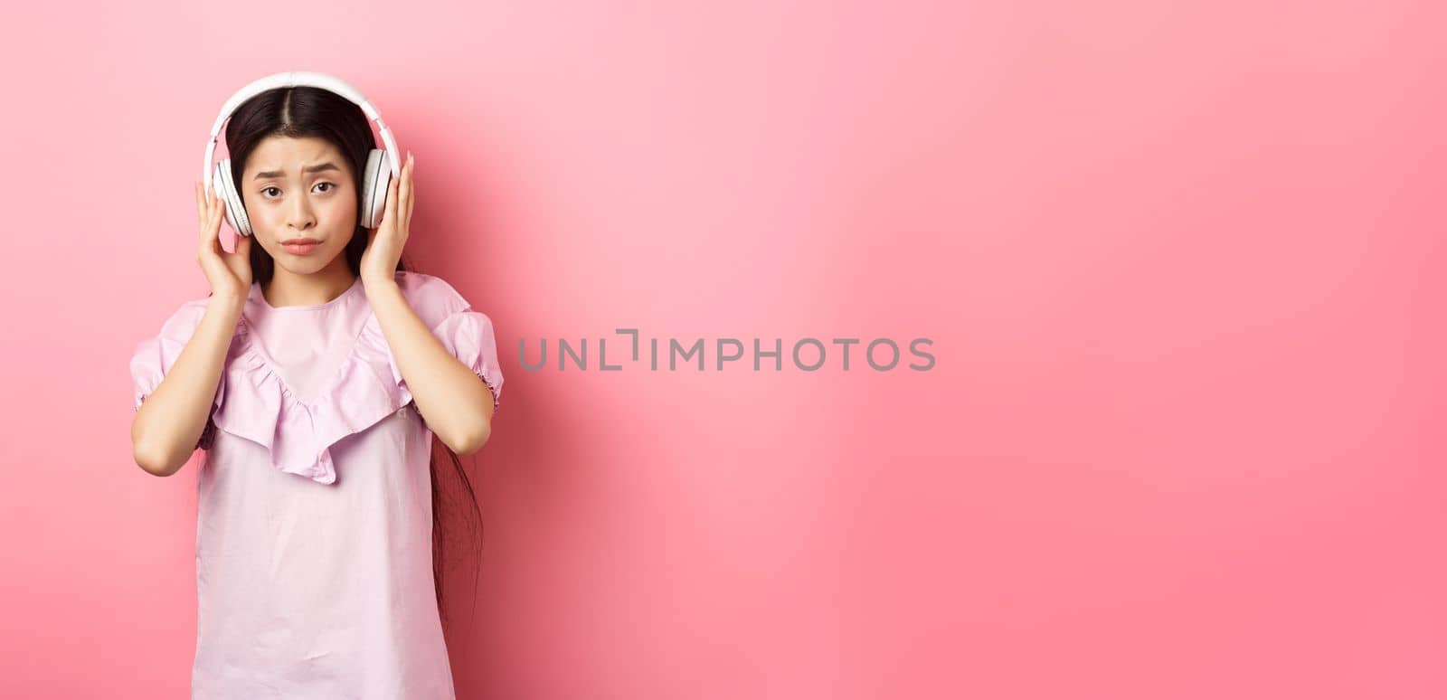 Skeptical girl unamused with song, listening music in headphones and frowning displeased, standing against pink background.