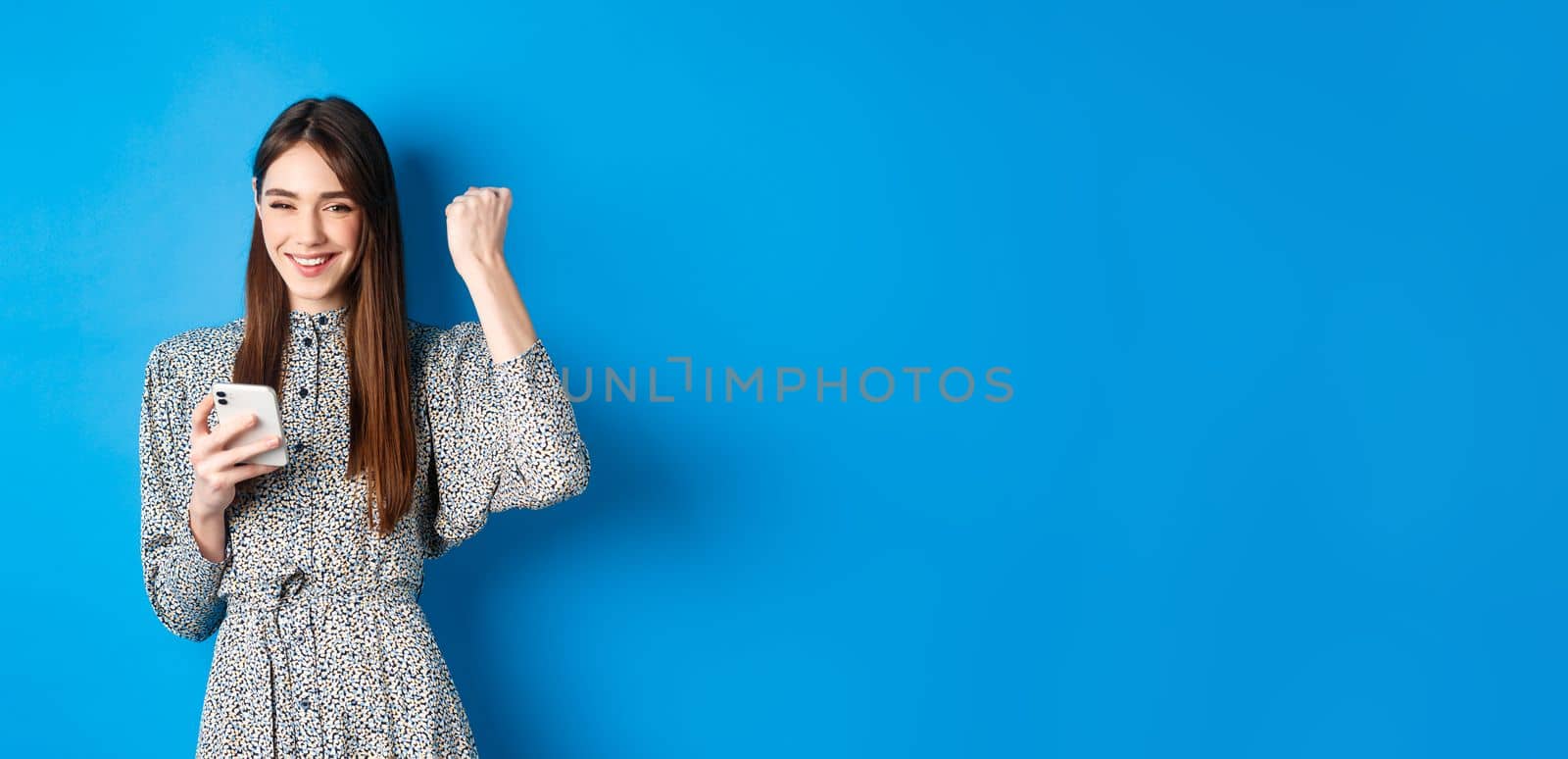 Happy pretty girl winning on mobile phone, smiling and chanting, holding smartphone, standing in dress against blue background.
