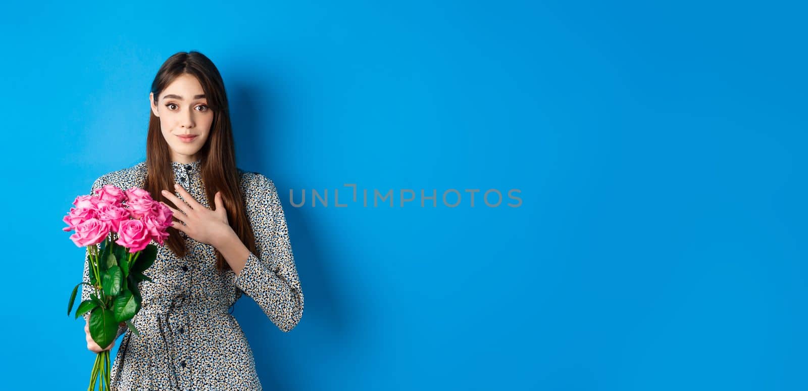 Valentines day concept. Touched and thankful pretty girl, holding hand on heart and smiling grateful for gift, holding bouquet of roses, standing on blue background.