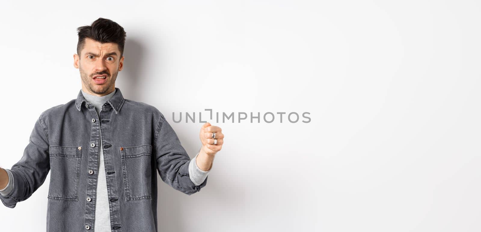 Skeptical young man showing big size object and frowning doubtful, standing reluctant on white background.