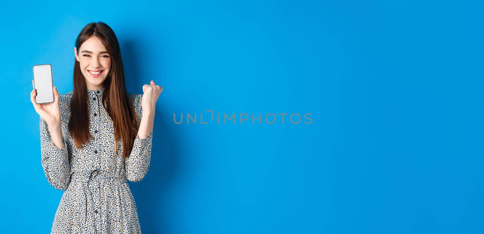 Cheerful pretty girl chanting, winning on mobile phone, showing empty smartphone screen and fist pump, smiling and celebrating, blue background.