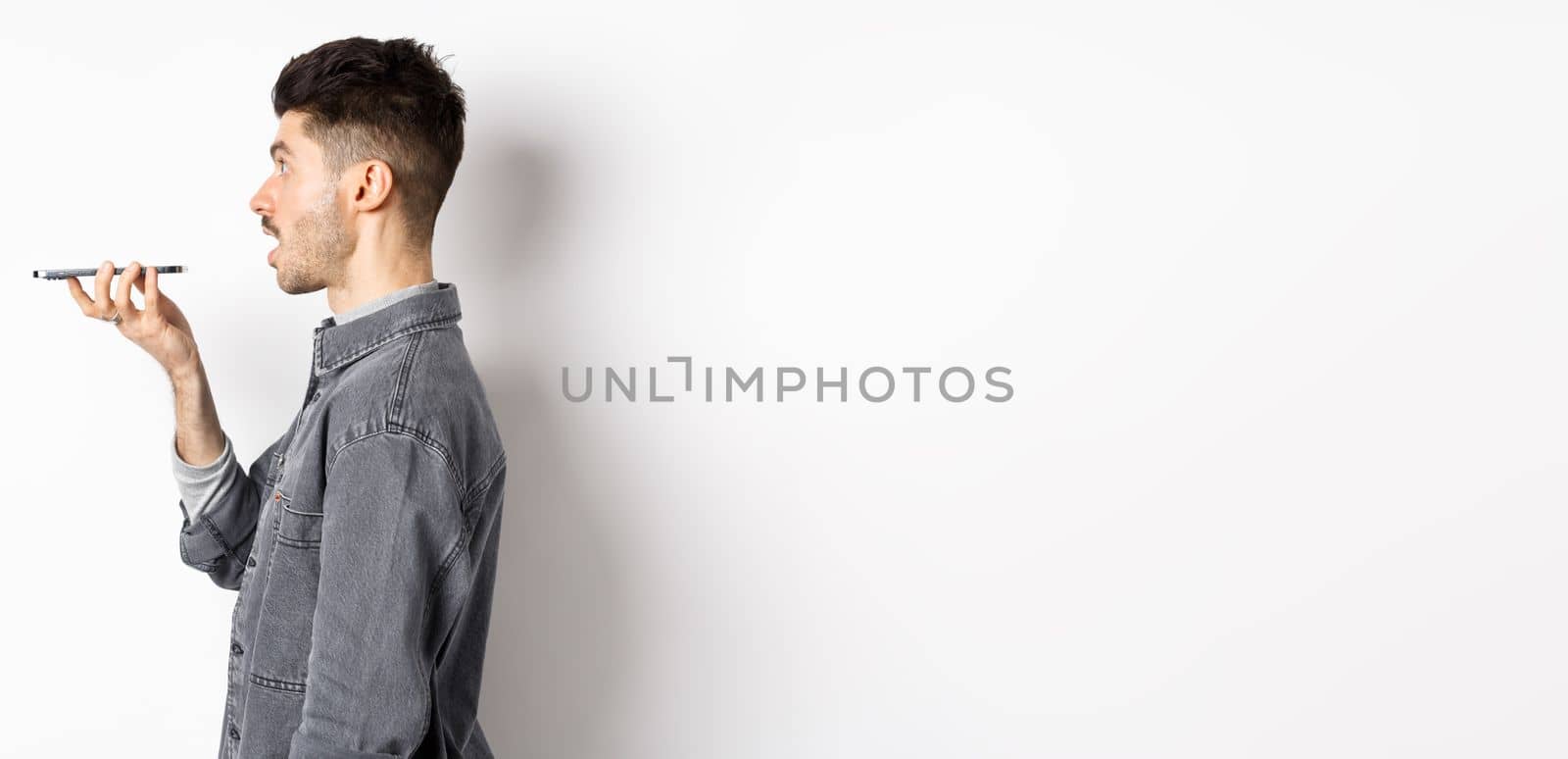 Profile of young man speaking at smartphone, translate with voice-translator app, talking into mobile phone speakerphone, standing on white background.