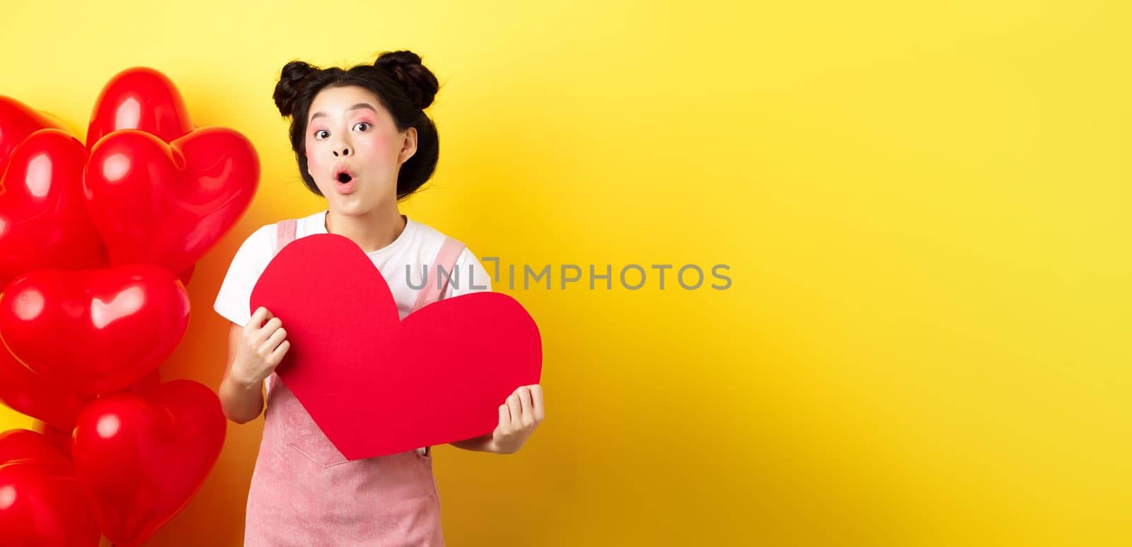 Happy Valentines day. Silly asian girl showing big red heart postcard and looking amazed, standing near red romantic balloons, yellow background.