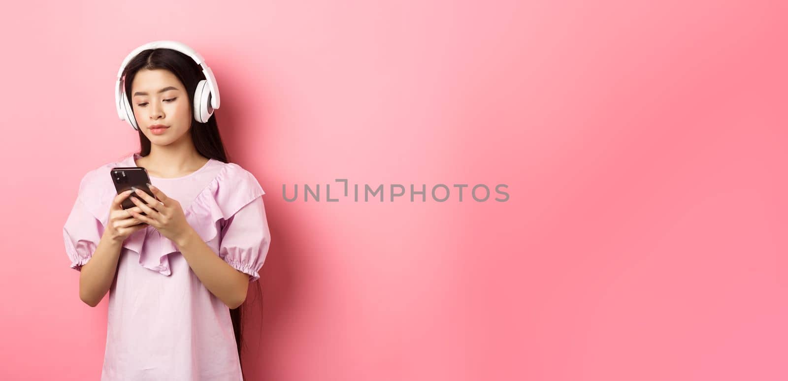 Young girl listening music in headphones and chatting on mobile phone, standing in dress against pink background.