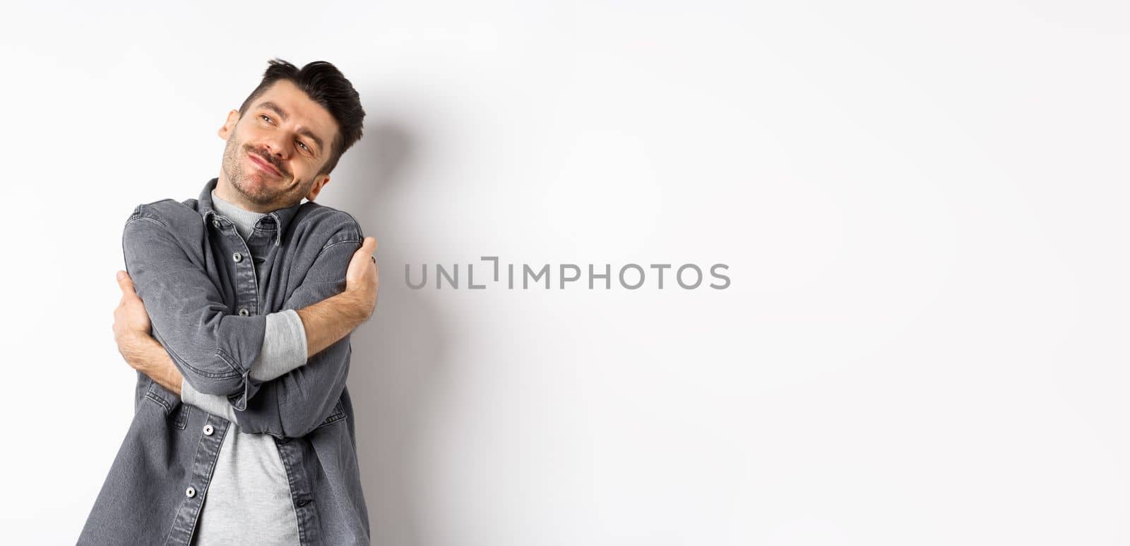 Happy romantic guy hugging himself with closed eyes and smile, loving own body, daydreaming about lover, standing on white background by Benzoix