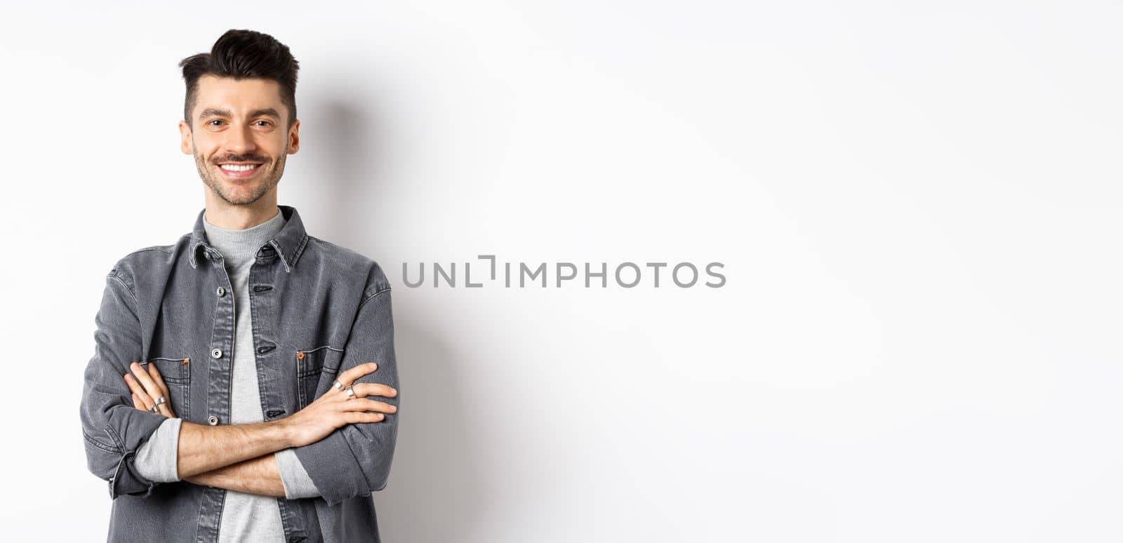 Handsome young man smiling with arms crossed on chest, feeling ready, looking motivated at camera, standing on white background.
