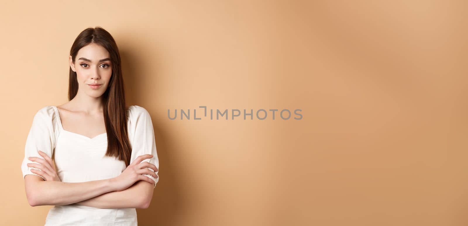 Young caucasian woman looking determined and confident, standing with arms crossed, smiling at camera, wearing white dress, beige background.