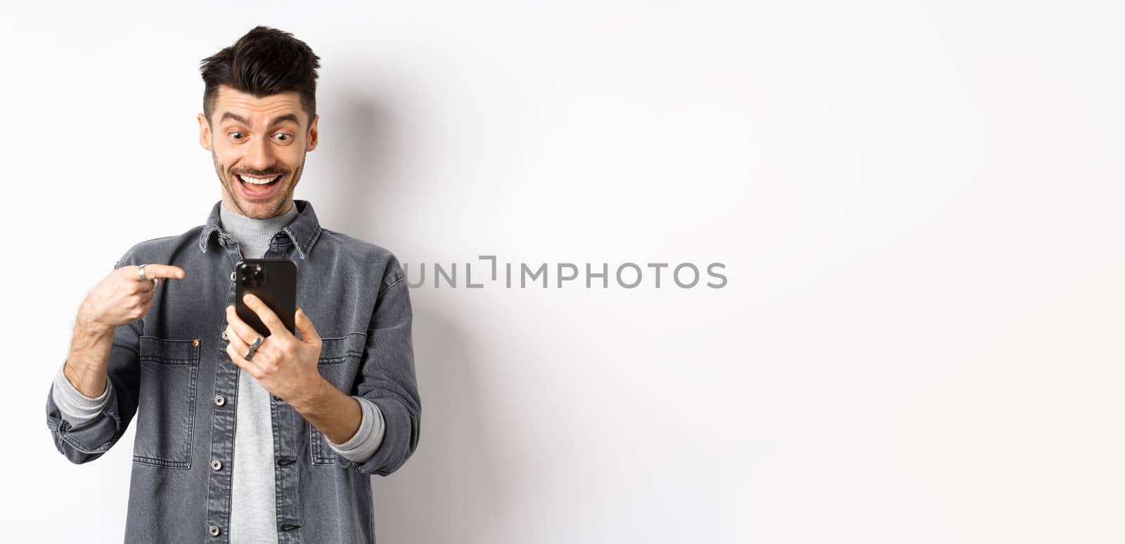 Excited handsome man showing cool news on phone, pointing at smartphone and smiling happy, standing against white background.