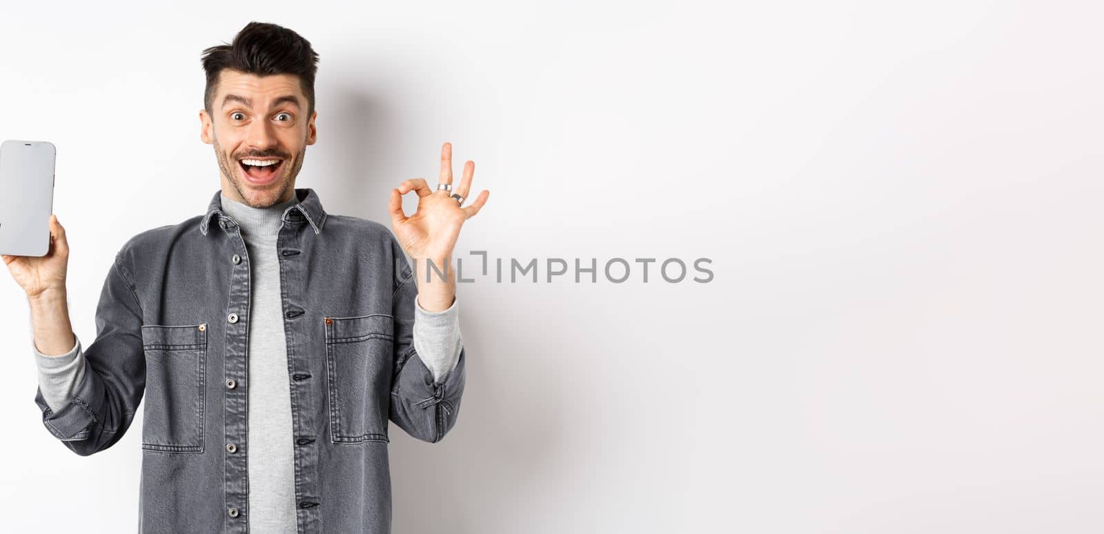 Excited smiling guy showing empty smartphone screen with okay gesture, recommending app or shopping offer, standing on white background.