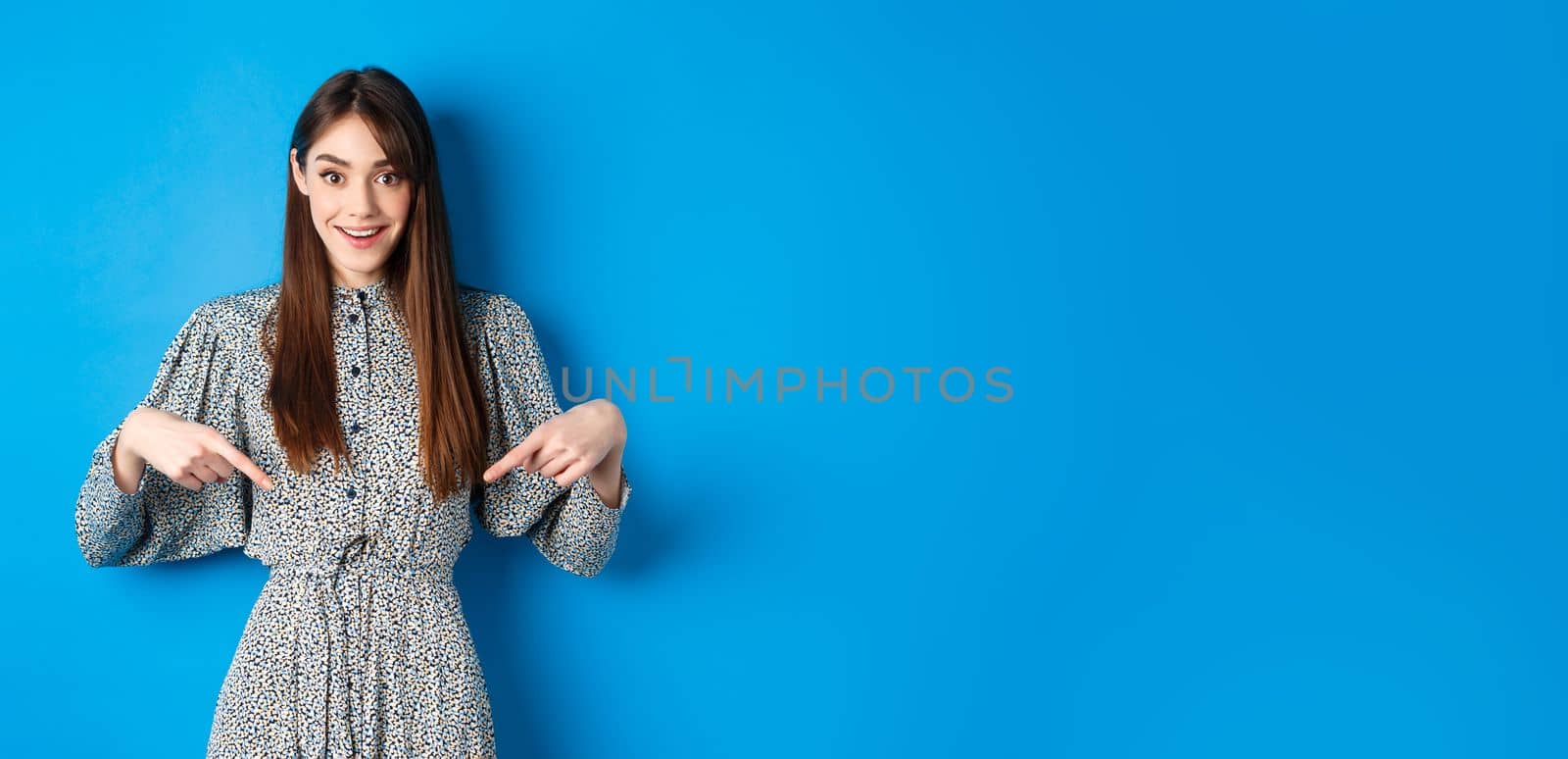 Excited caucasian woman in dress pointing fingers down at logo, showing fantastic news and smiling, standing on blue background by Benzoix