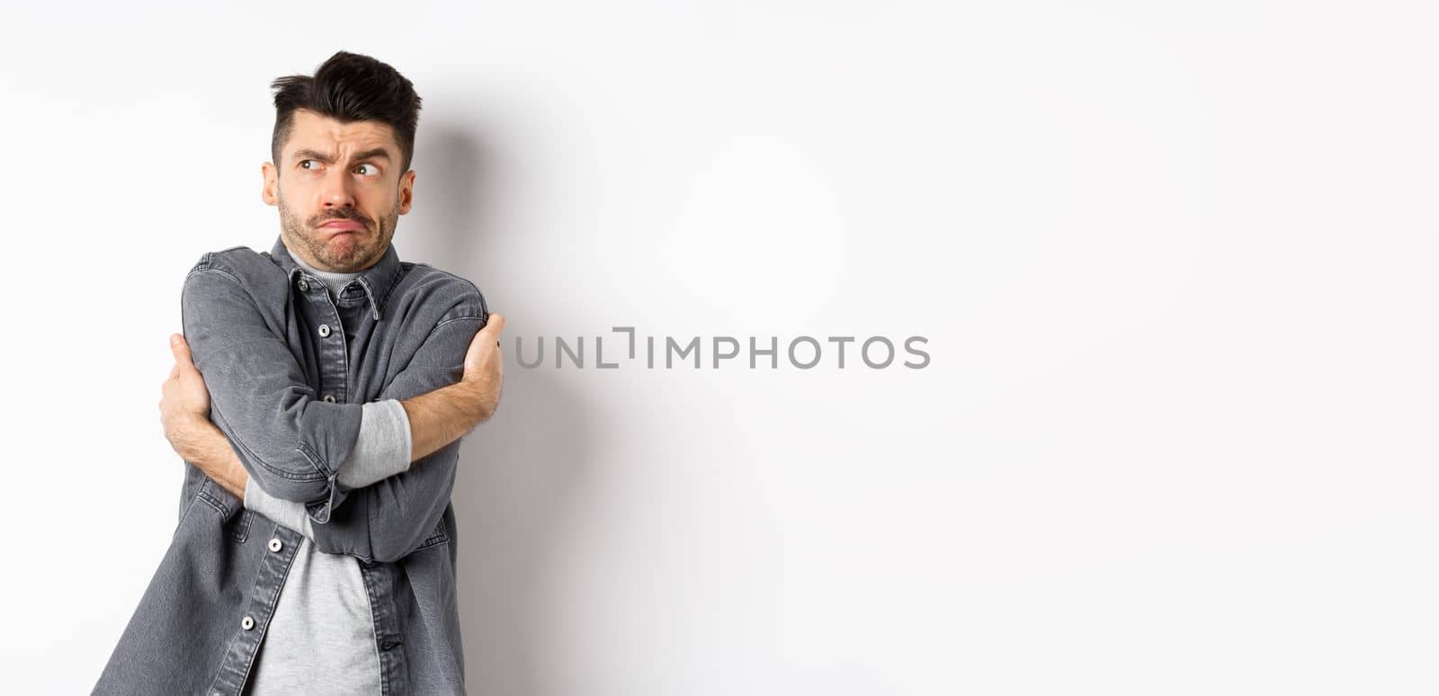 Displeased young man feeling discomfort, hugging himself and staring left at something embarrassing or exposing, standing uncomfortable on white background.