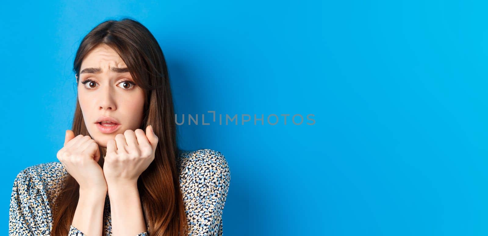 Close-up of scared timid girl looking afraid, shaking from fear, press hands to body and staring frightened, standing on blue background by Benzoix