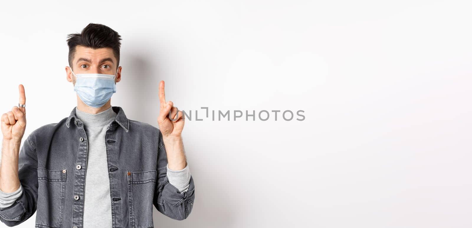 Pandemic lifestyle, healthcare and medicine concept. Stylish modern man in medical mask showing advertisement, pointing fingers up at logo, standing on white background.