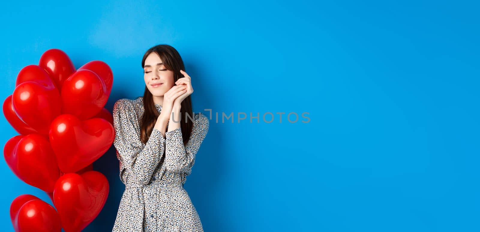 Valentines day. Dreamy romantic woman close eyes and imaging lovely date, standing near heart balloons and smiling, blue background by Benzoix