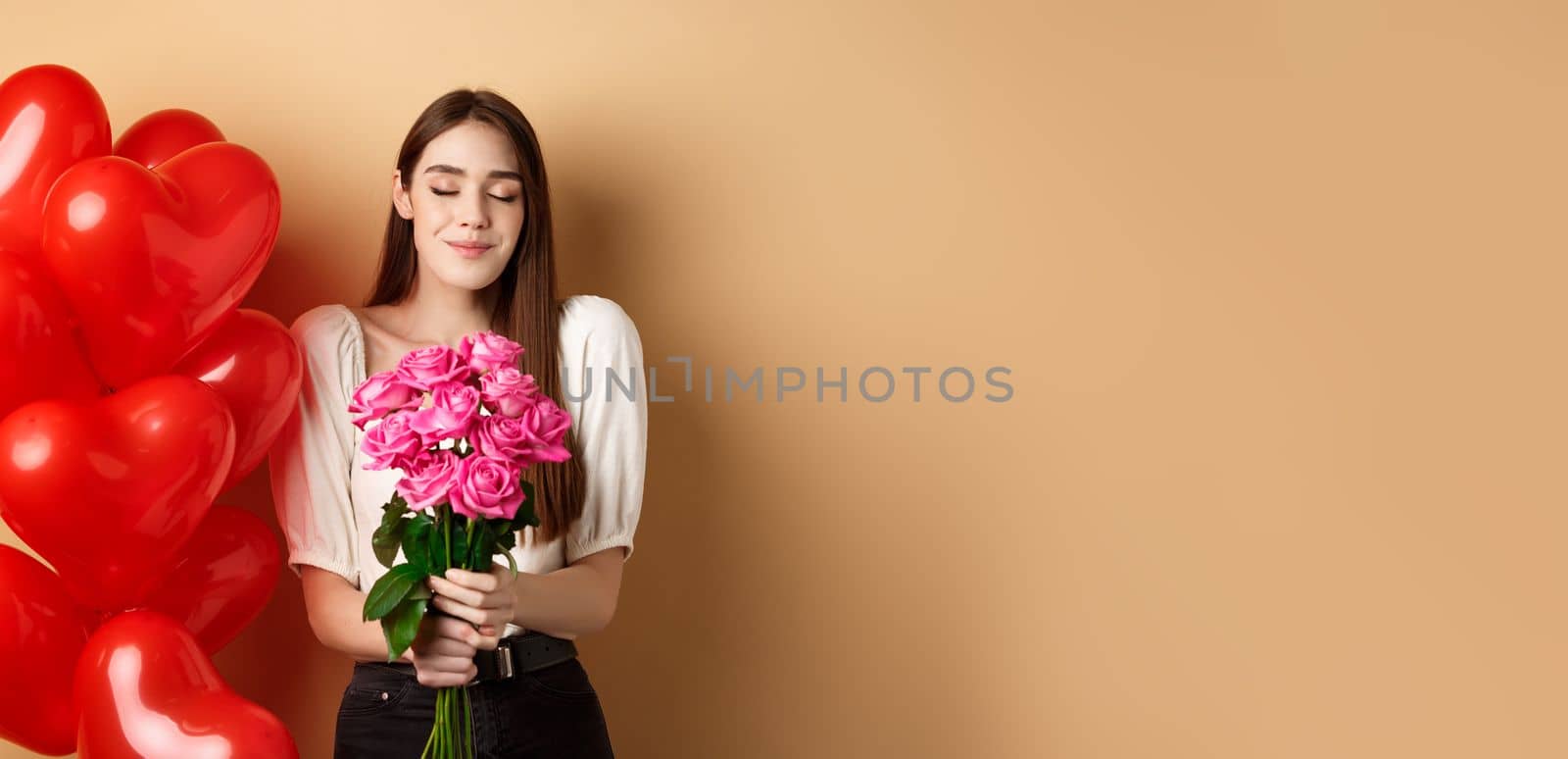 Romantic girlfriend smelling roses, receive flowers from lover on Valentines day, standing near red hearts balloons and smililng, beige background.