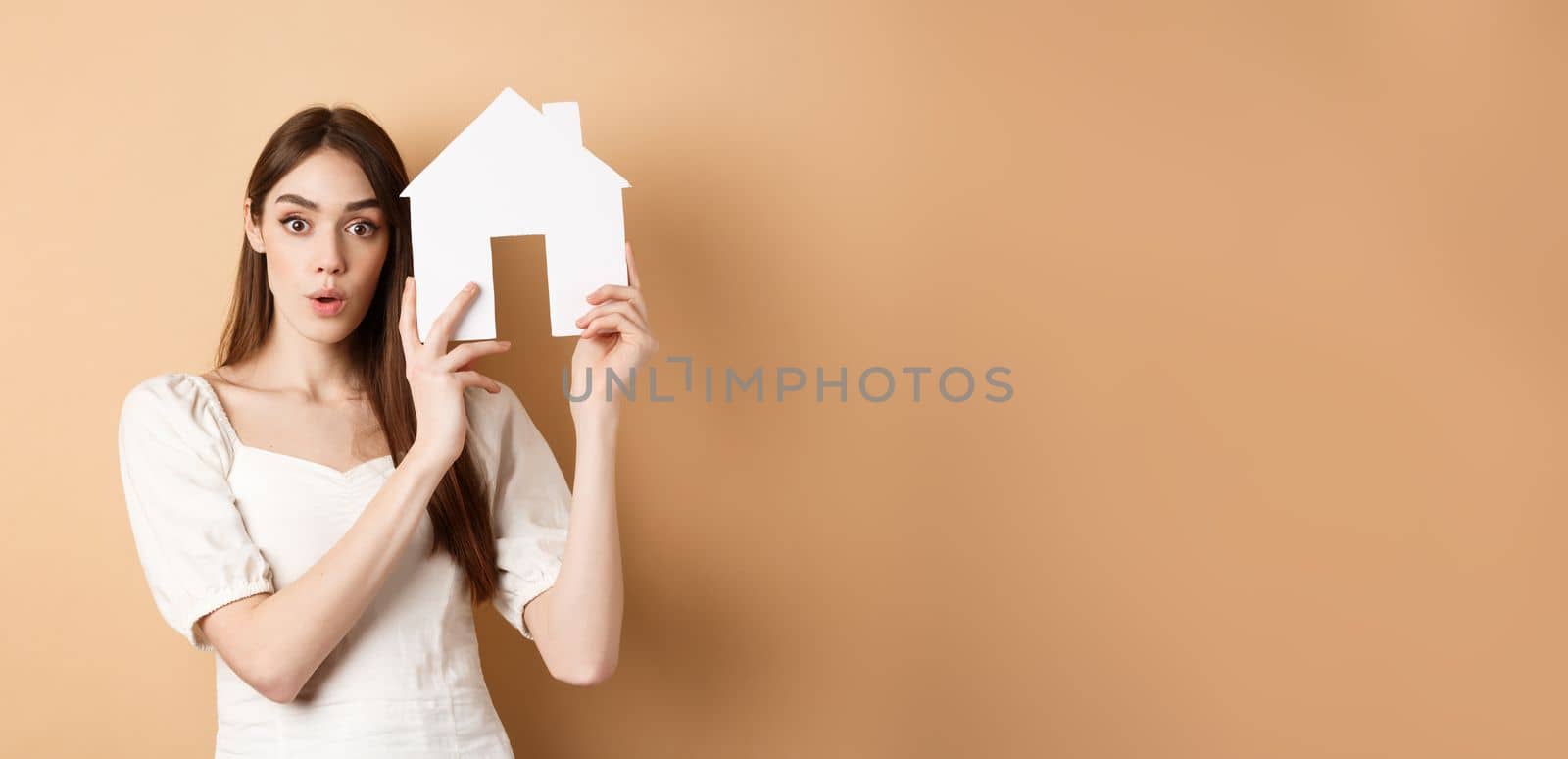 Real estate. Excited young woman showing house cutout and looking at camera, renting property, standing on beige background.