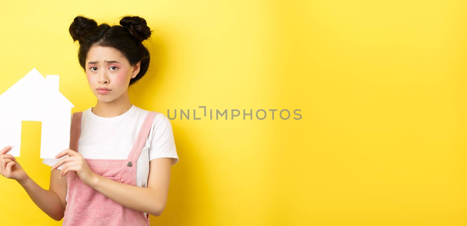 Real estate and family concept. Sad cute asian girl with bright makeup, frowning and feel upset, showing paper house cutout, standing on yellow background.