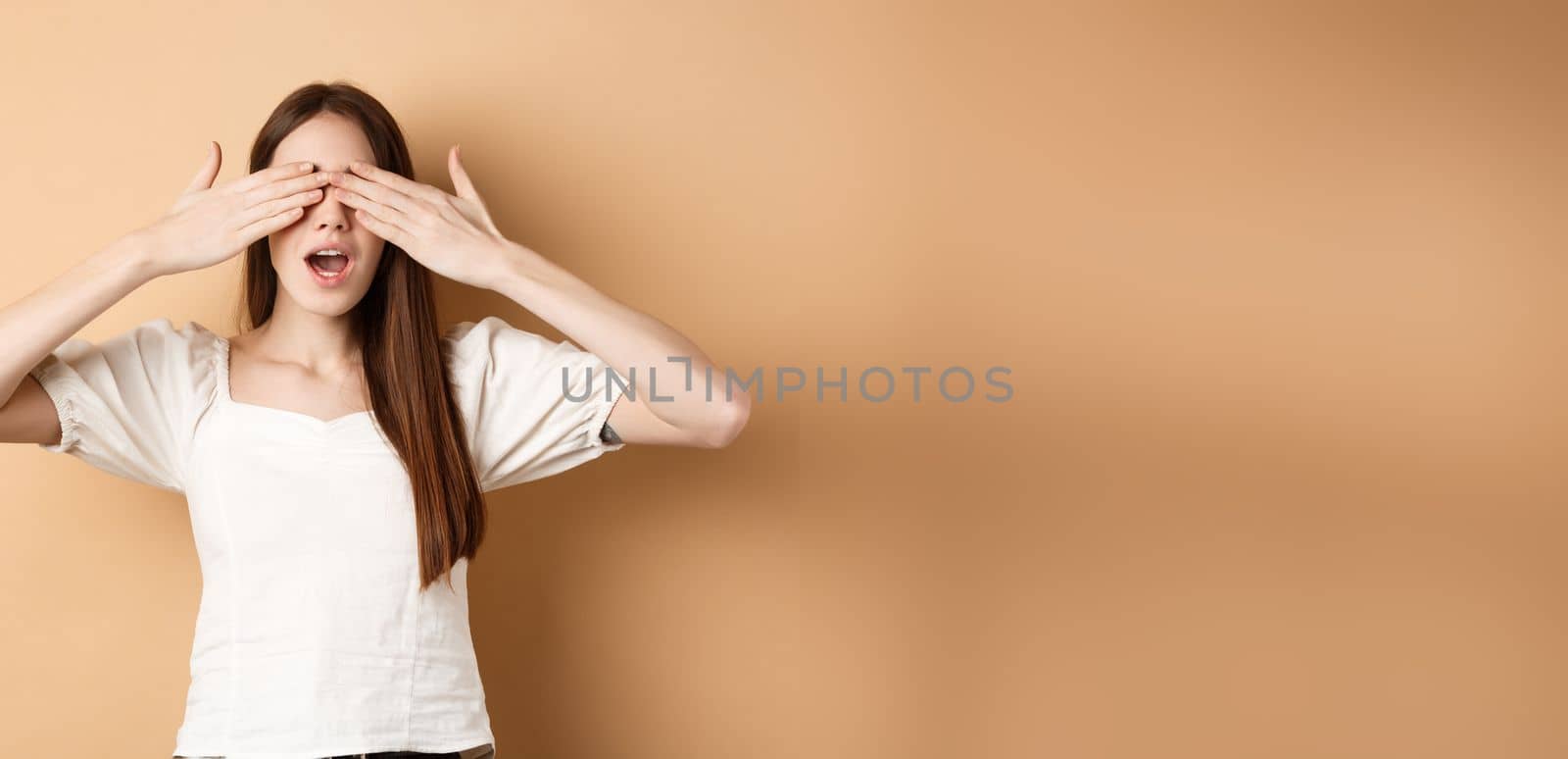 Happy romantic girl covering eyes and waiting for surprise, smiling while anticipating gift, standing on beige background.