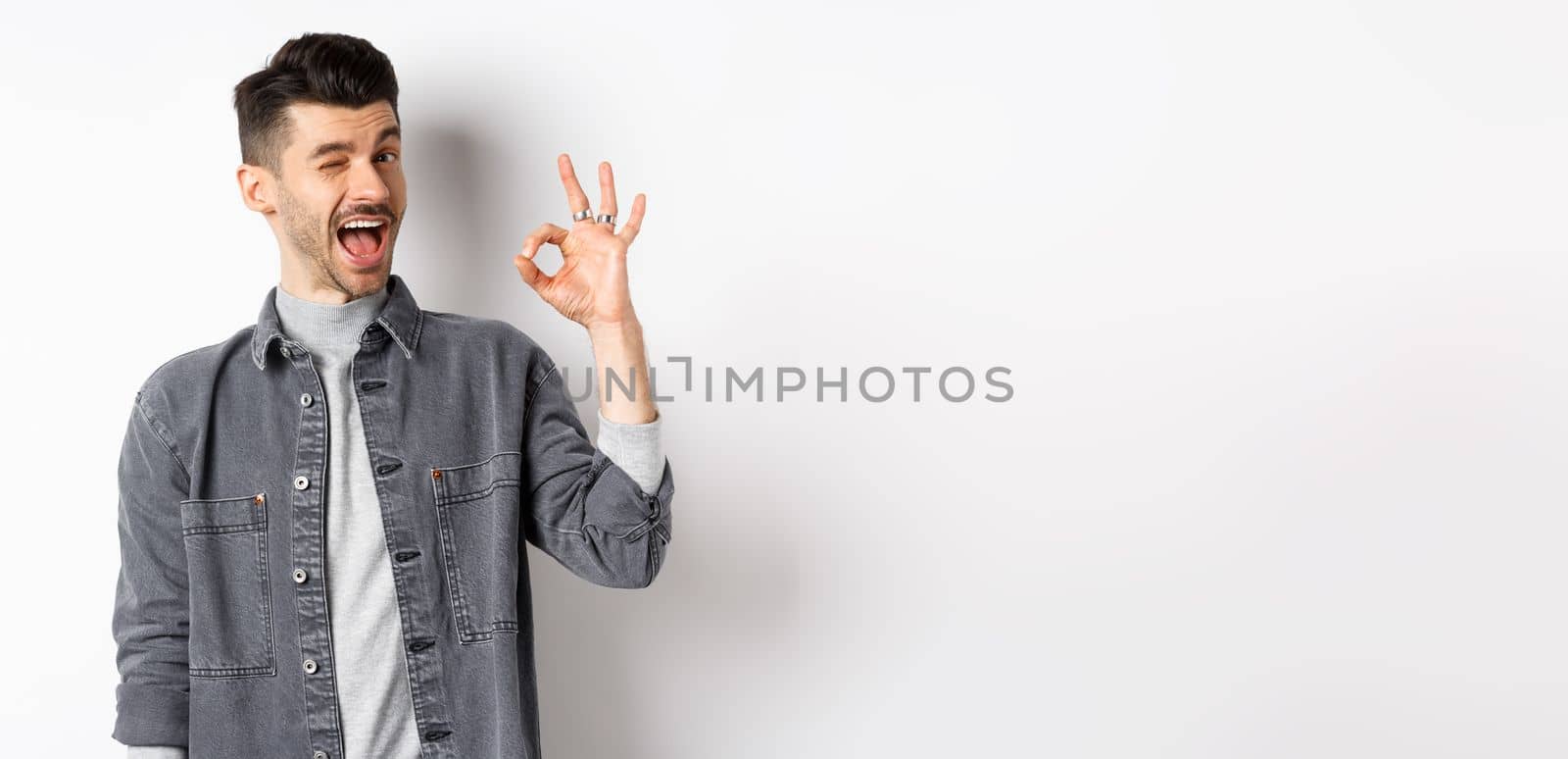 Excited handsome guy with moustache, winking and showing okay sign, smiling pleased, assure all good, praise nice work, making good job gesture, standing against white background.