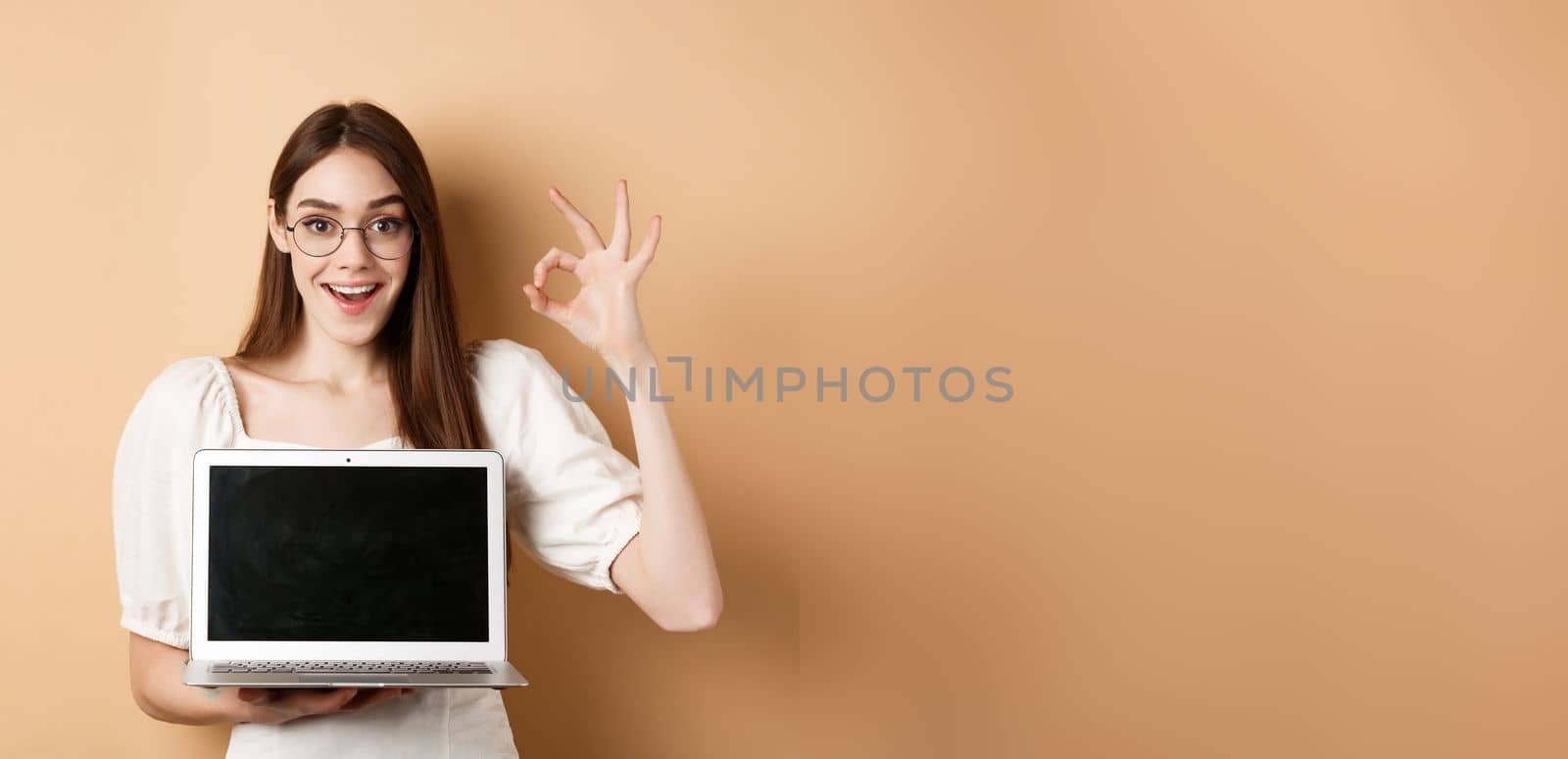 E-commerce. Excited young woman in glasses showing okay sign and laptop screen, recommending internet promo, standing on beige background.