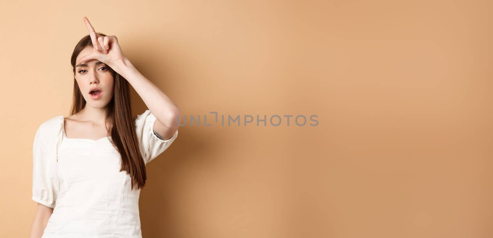 Confident woman mocking people with loser sign, being mean, standing on beige background.