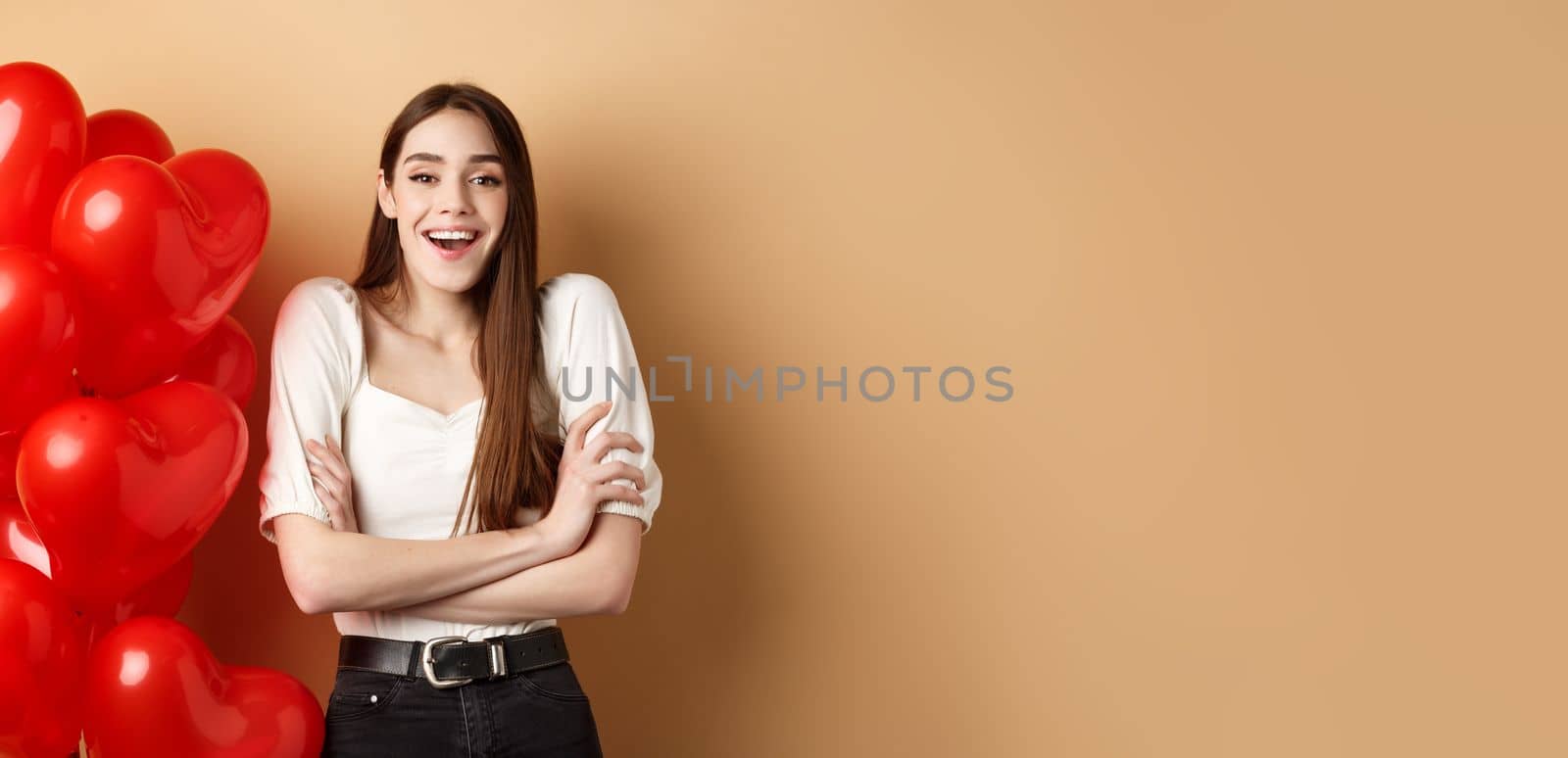 Valentines day concept. Beautiful young woman having fun, laughing and smiling at camera, standing near romantic heart balloons, beige background.