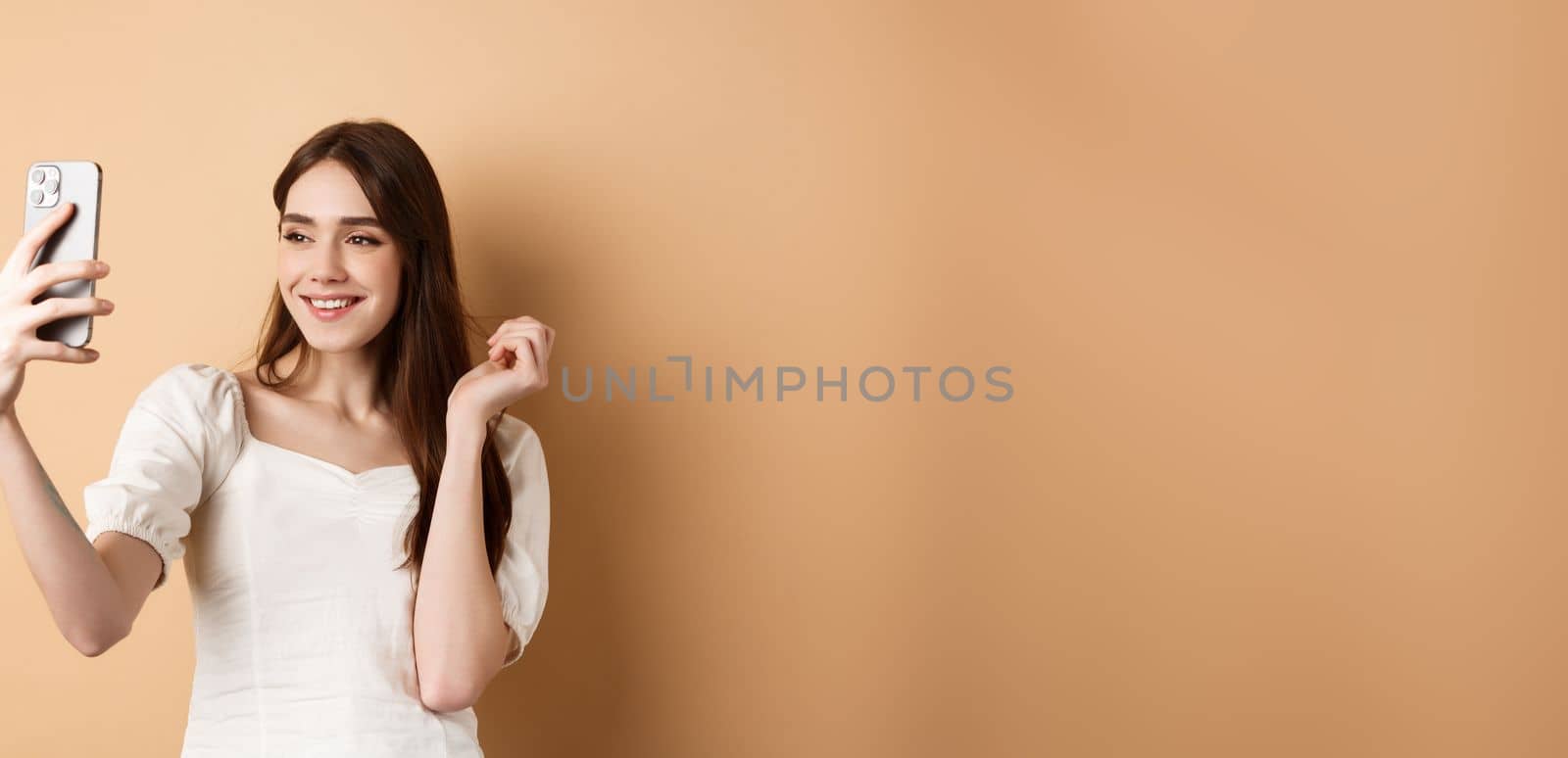 Beautiful woman taking selfie on smartphone, posing for phone photo, standing on beige background.