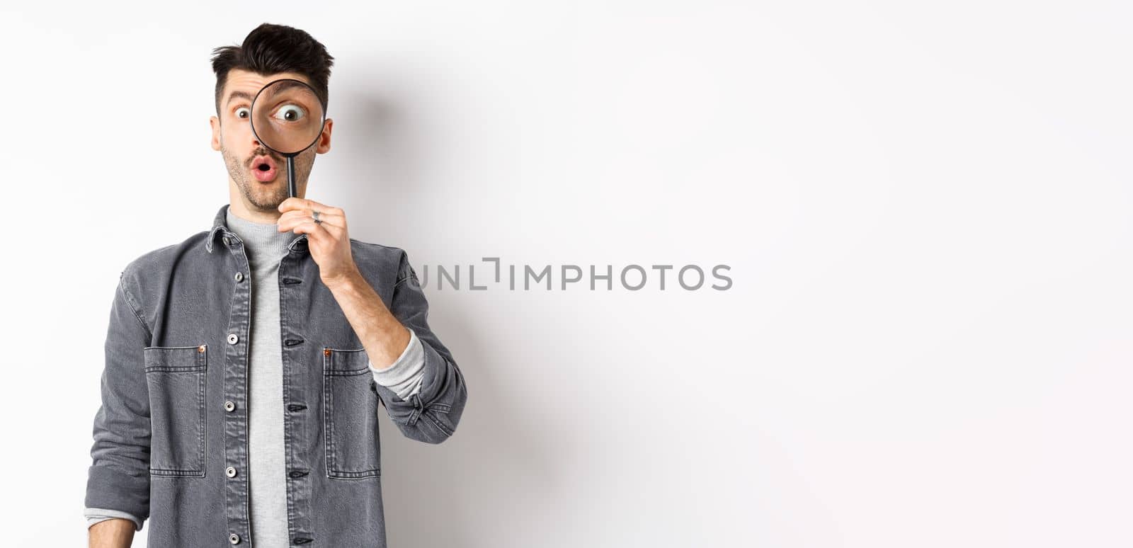 Image of excited guy look through magnifying glass, gasping amazed, big eye staring at interesting thing, found cool promo, standing on white background.