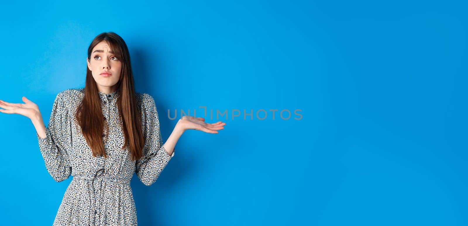 Confused pretty girl in dress shrugging shoulders and looking aside sad, know nothing, standing against blue background.