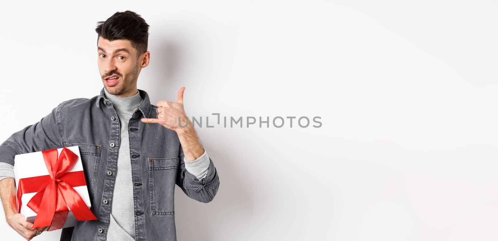 Macho guy with gift box show call me phone gesture, flirting with you, standing boastful on white background. Relationship and Valentines day concept by Benzoix