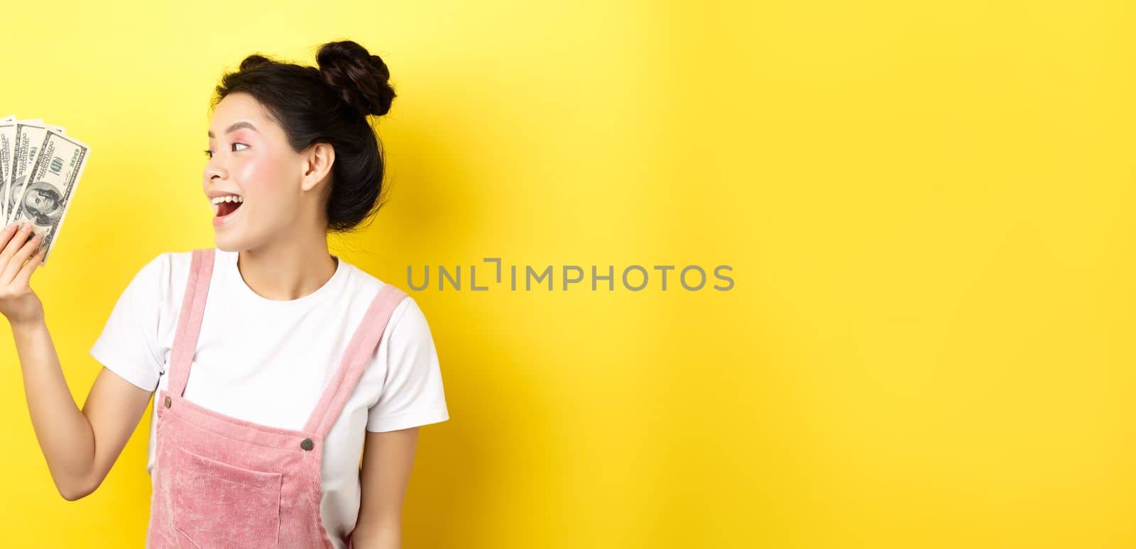 Shopping. Rich and stylish asian female model showing money, looking at dollar bills with happy face, standing on yellow background by Benzoix