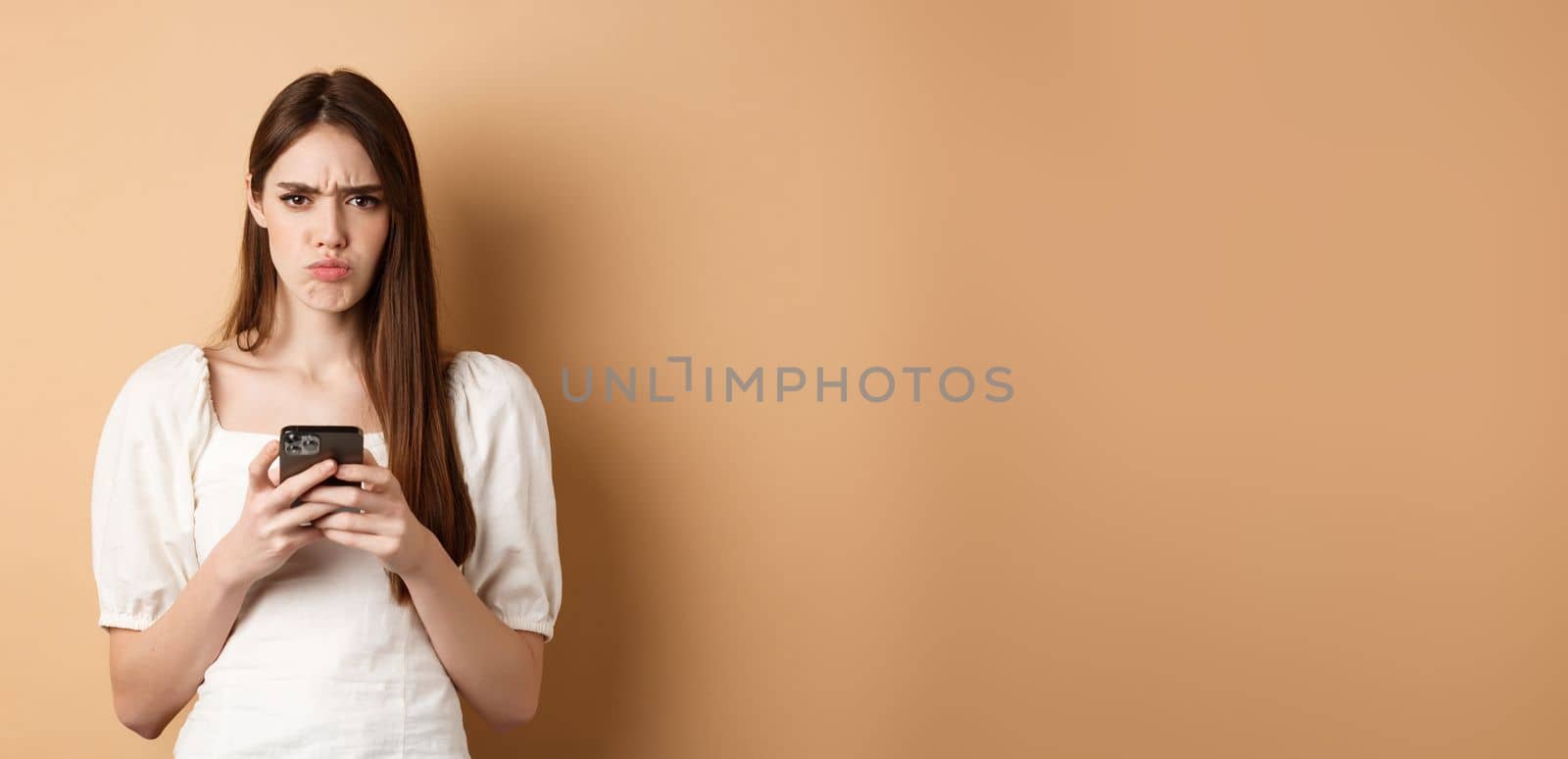 Disappointed girl with smartphone frowning, pucker lips upset, reading bad news on phone, standing on beige background.