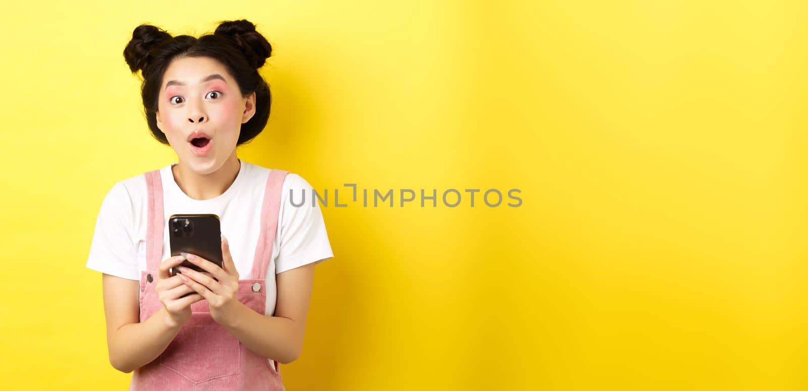 Excited girl with glam makeup and summer clothes, gasping amazed, checking out internet offer on mobile phone, standing on yellow background.