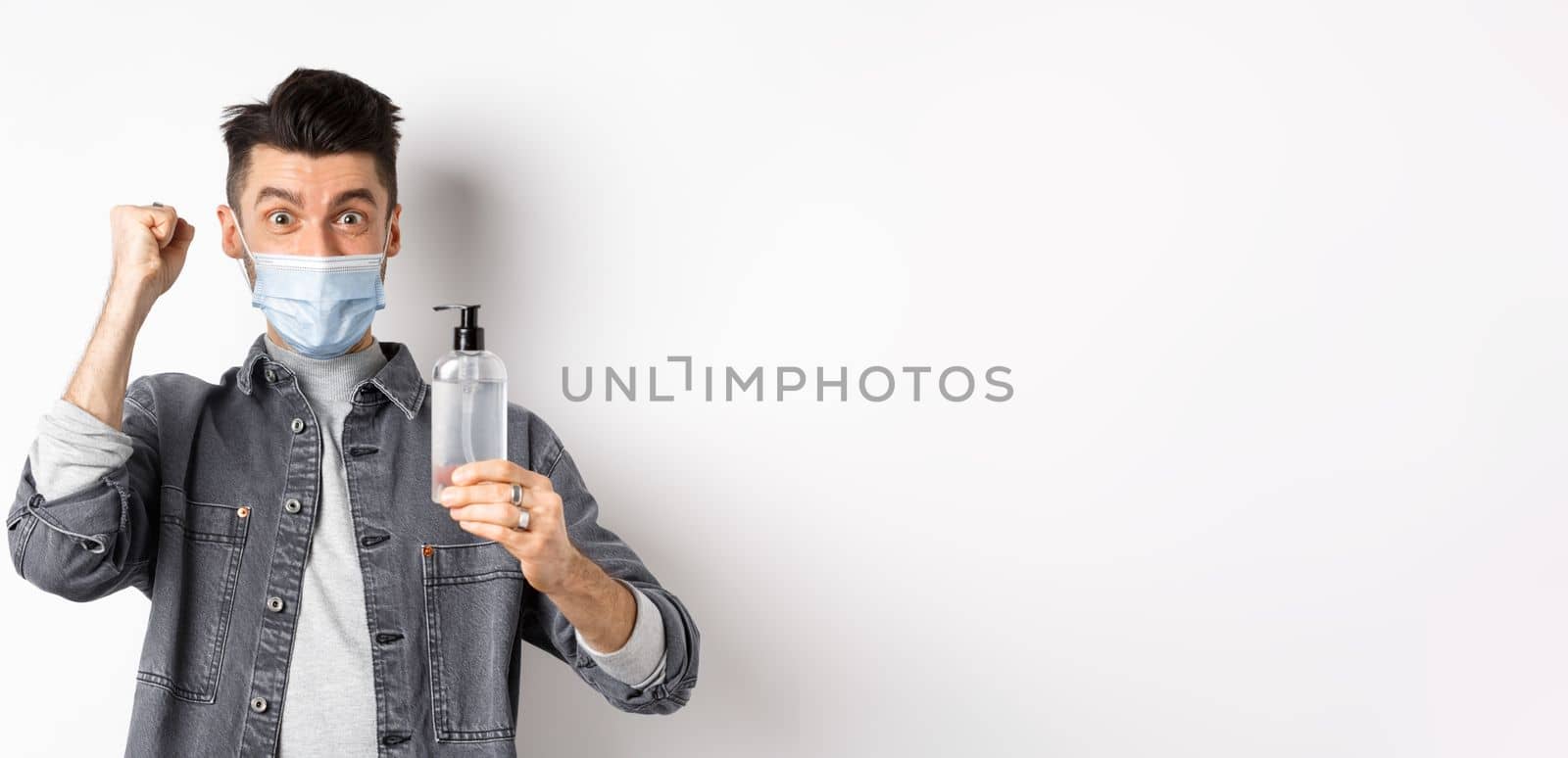 Health, covid and pandemic concept. Happy guy motivated to get rid from virus germs, showing bottle of hand sanitizer and fist pump, cheering while standing on white background.