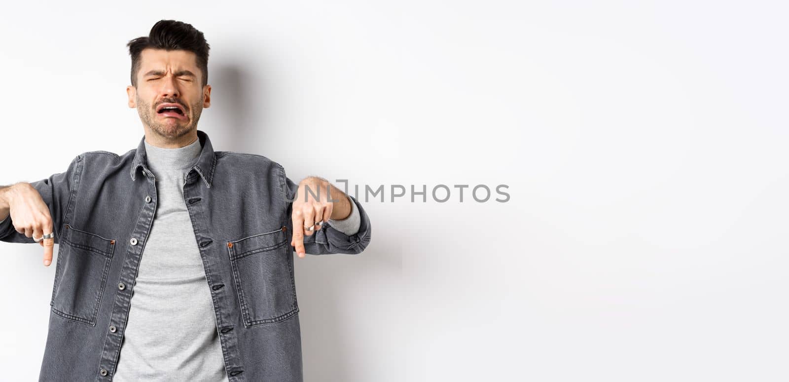 Sad crying man pointing fingers down, sobbing miserable, showing upsetting news, standing in denim jacket on white background.