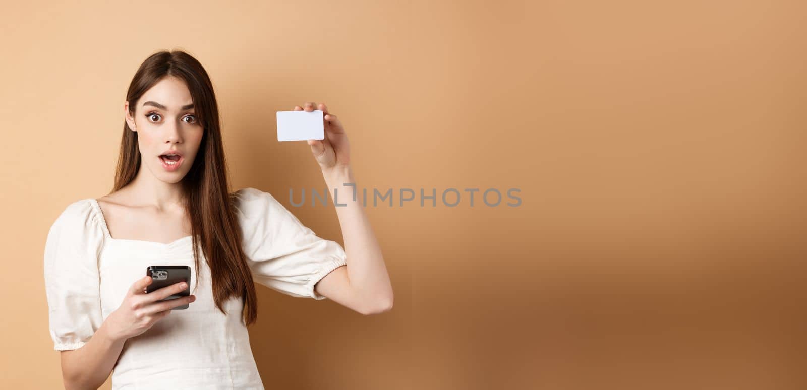 Excited woman showing plastic credit card and using mobile phone app, drop jaw and gasping amazed, checking out bank offer, beige background.