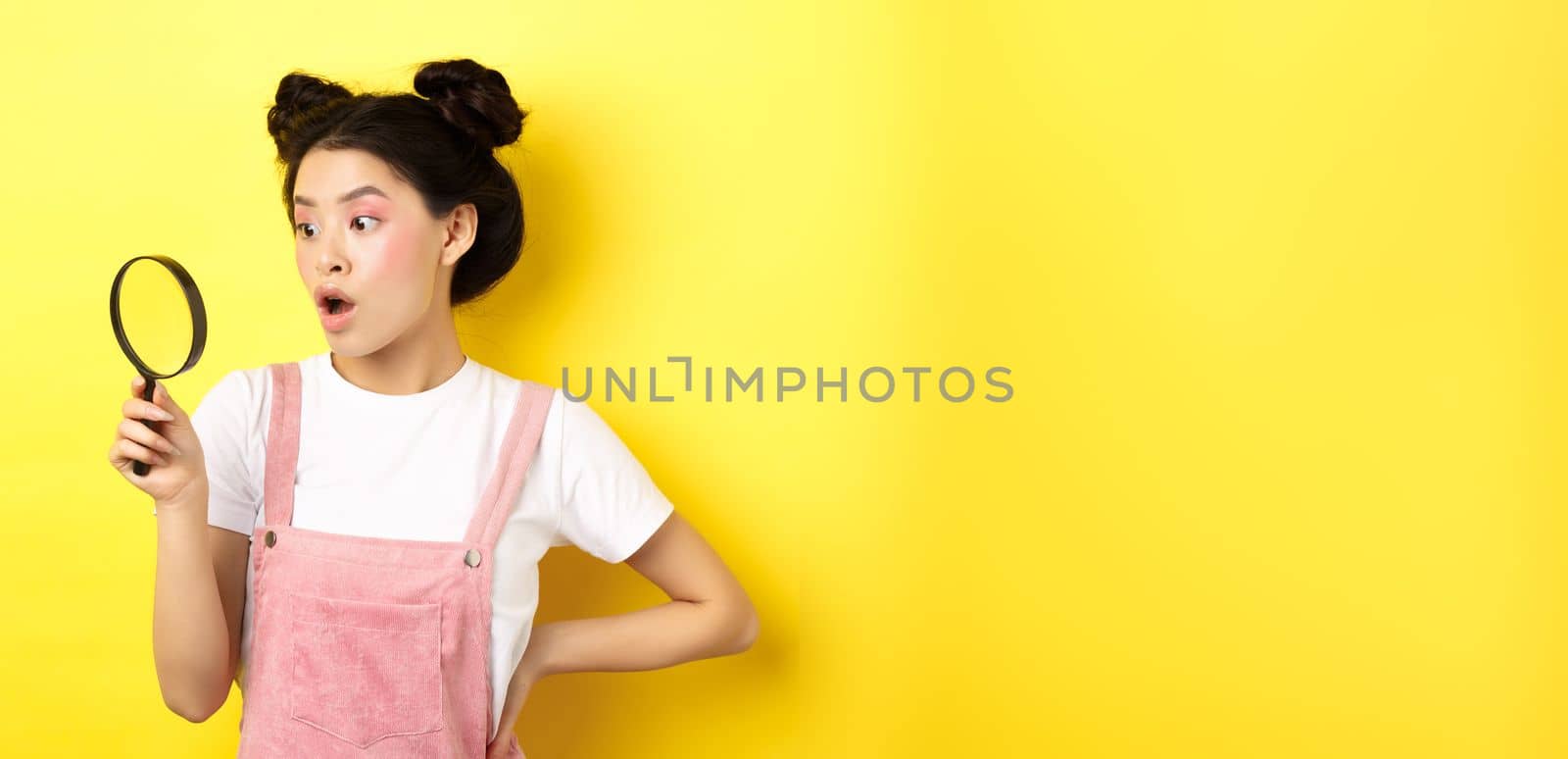 Excited asian woman with bright glamour makeup, look through magnifying glass amazed, staring aside, standing on yellow background.