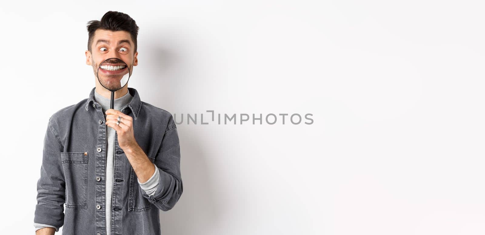 Handsome positive guy showing white perfect smile with magnifying glass, and squinting eyes, making funny faces, white background.