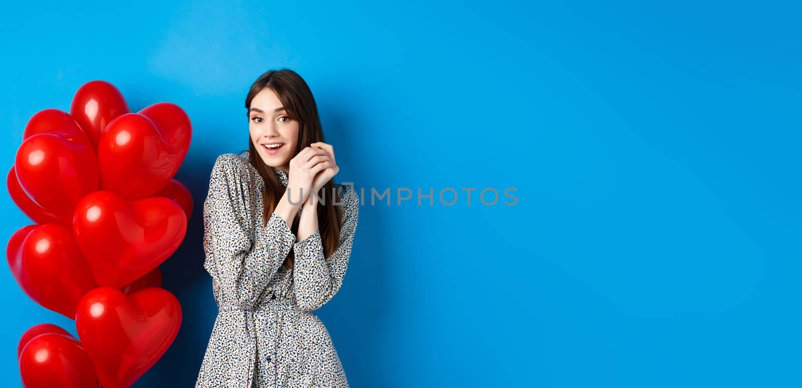 Valentines day. Lovely girlfriend ind ress smiling happy at camera, celebrating lovers holiday, standing near heart balloons, blue background by Benzoix