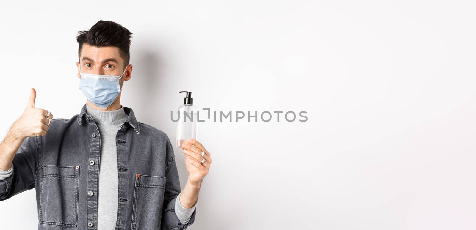 Healthy people and covid-19 concept. Excited man in sterile medical mask holding bottle of good hand sanitizer, show thumb up, recommend antiseptic, standing against white background.