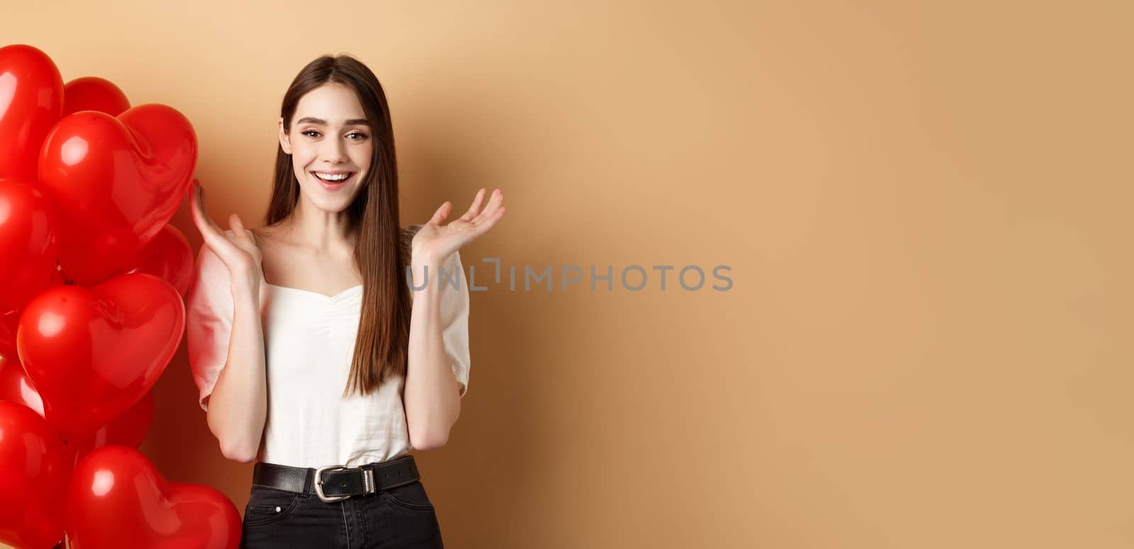 Valentines day and love concept. Happy smiling woman raising hands up surprised, standing near romantic heart balloons on beige background by Benzoix