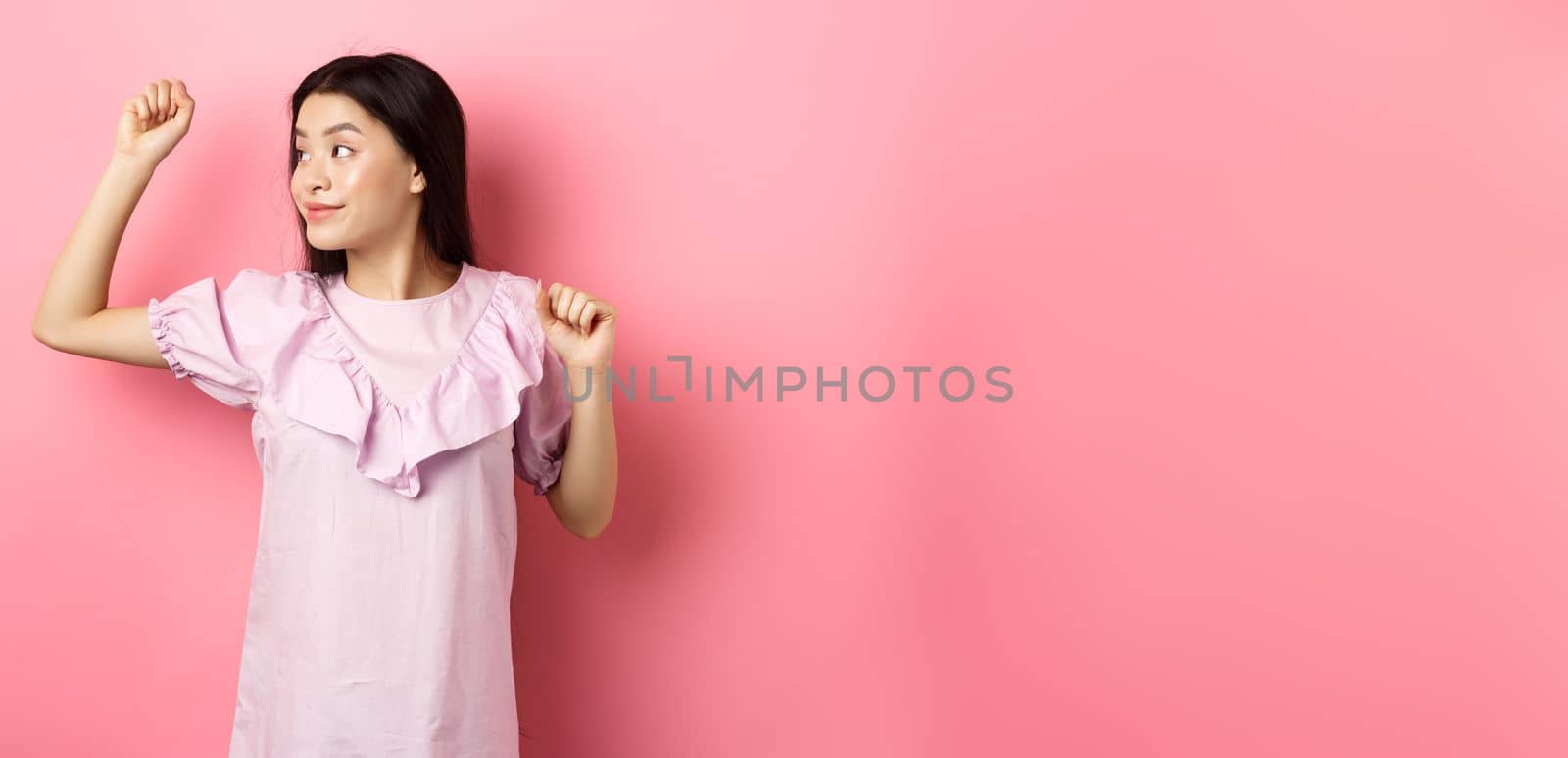 Carefree asian girl dancing, raising hand up and looking left at logo, standing in romantic dress against pink background.