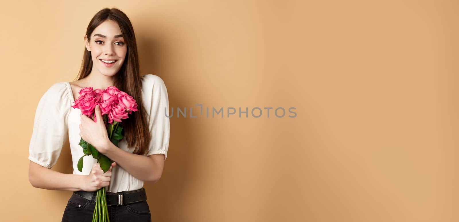 Valentines day. Romantic girl smiling happy at camera, holding bouquet of pink roses from lover, standing on beige background by Benzoix