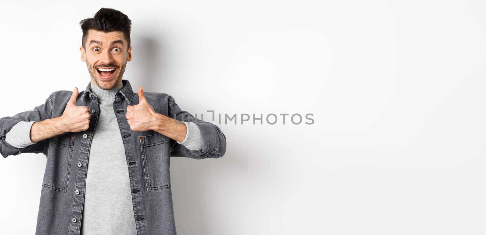 Sounds great. Excited young man showing thumbs up and smiling happy, recommending cool thing, praise nice job, standing on white background.